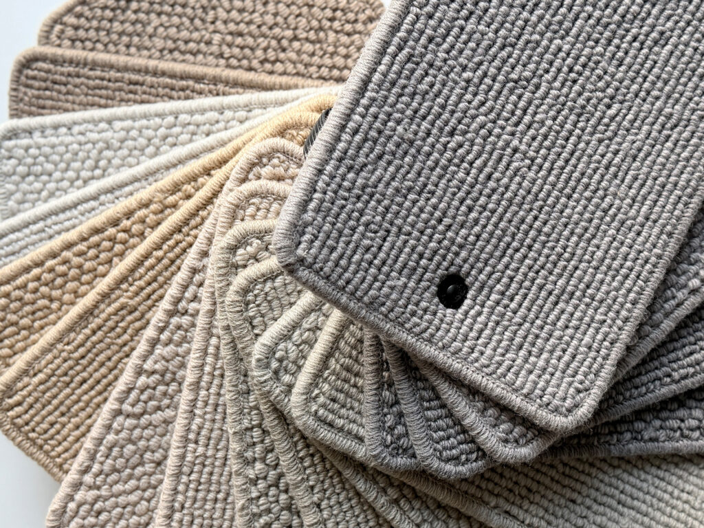 A fan of natural fibre carpet samples in soft neutral tones, ranging from light beige to warm grey. The close-up view highlights the durable woven textures and craftsmanship of wool and plant-based materials, illustrating the strength and resilience of natural fibres in sustainable flooring design.