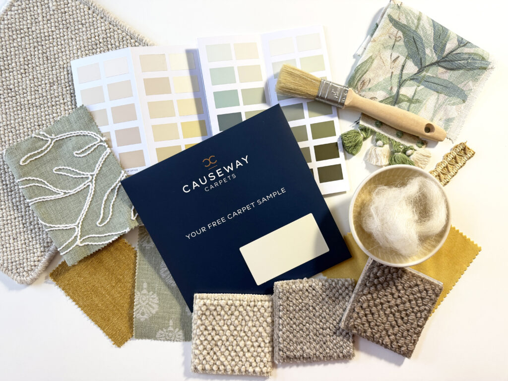 Flat lay of 100% wool carpet samples from Causeway Carpets, featuring textured loop piles in neutral tones (cream, beige, taupe, and brown), fabric swatches in mustard and sage patterns, green and neutral paint colour cards, a natural bristle paintbrush, yarn tassels, and a bowl of raw wool, arranged on a light surface to showcase coordinating interior design materials.
