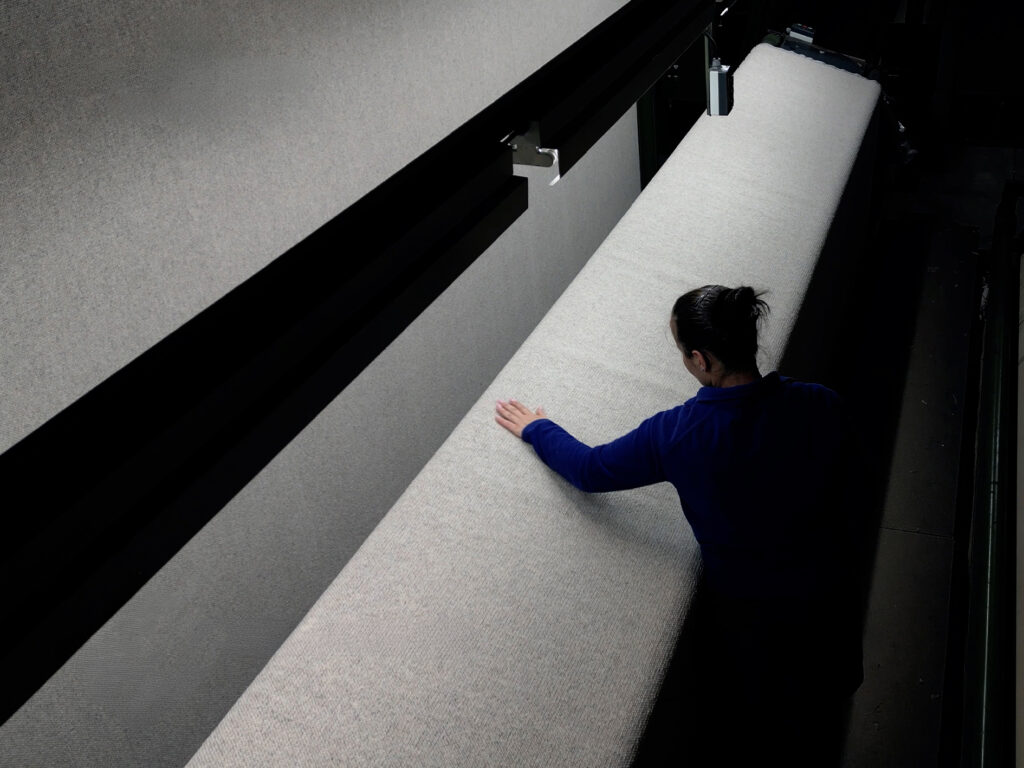 Person inspecting a large roll of light-colored carpet or fabric in a manufacturing or showroom setting.