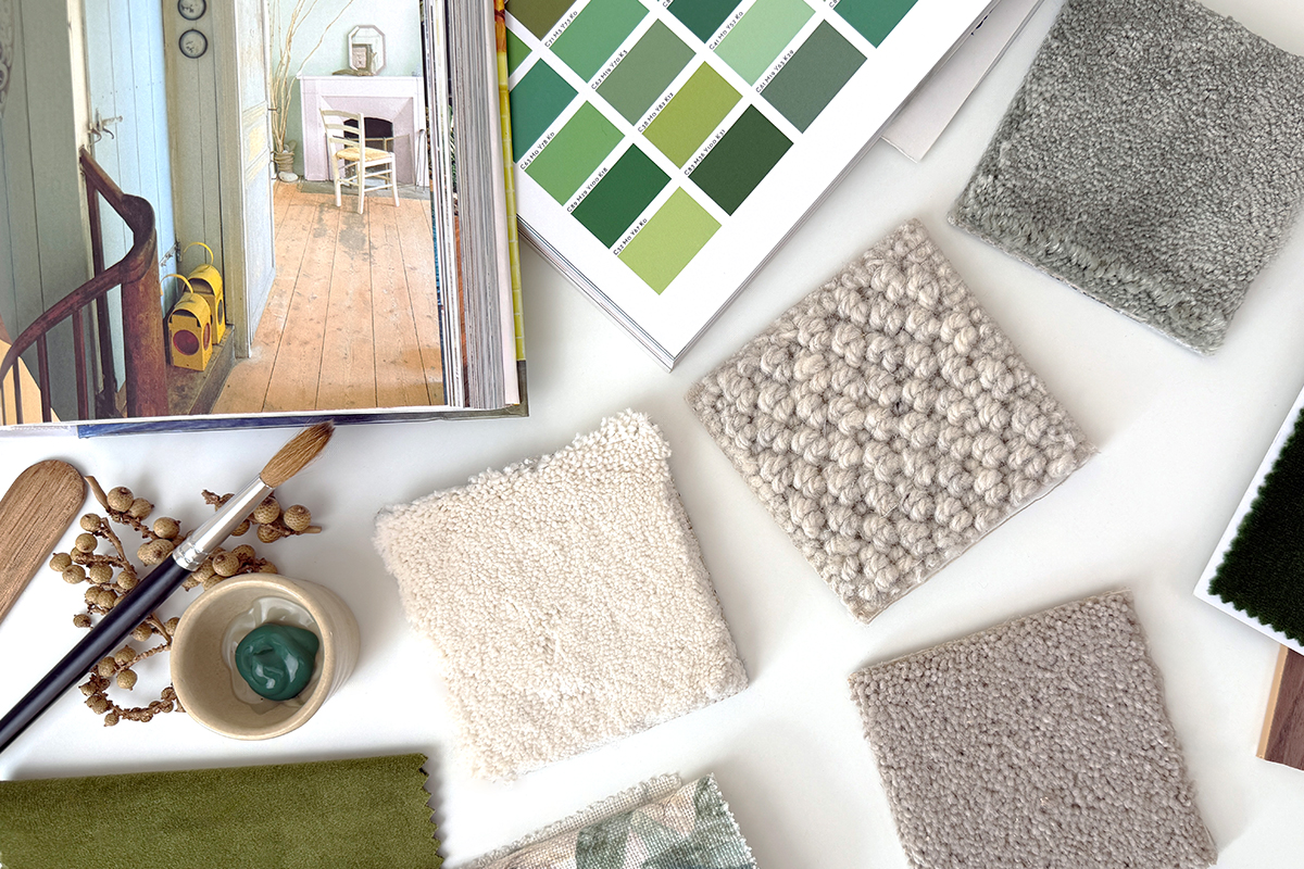 Interior design mood board featuring neutral carpet and fabric samples, green paint swatches, an open interiors book, a paintbrush, and a small dish of green paint arranged on a white surface.